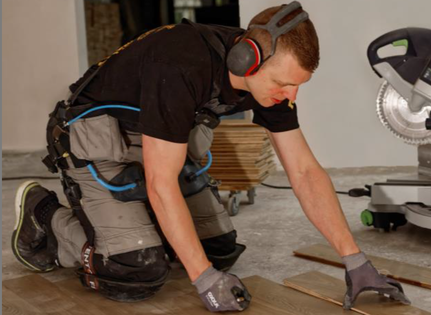 Ein Bodenleger trägt das Exoskelett und kniet arbeitend auf dem Boden.